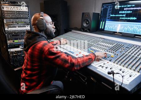 Stilvoller afroamerikanischer Musikproduzent, der Kopfhörer trägt und am Mischpult sitzt, um im Studio Songs aufzunehmen Stockfoto