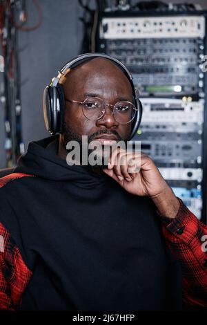 Porträt eines modernen afroamerikanischen Mannes mit Bart im Gesicht, der Kopfhörer und Brillen trägt und im Aufnahmestudio sitzt und die Kamera anschaut Stockfoto