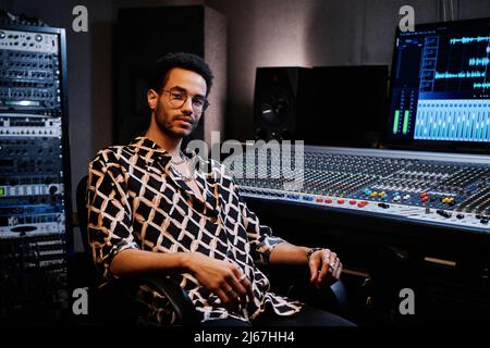 Der junge afroamerikanische Produzent trägt ein modisches Outfit und sitzt im Studio gegen eine Mischkonsole Stockfoto