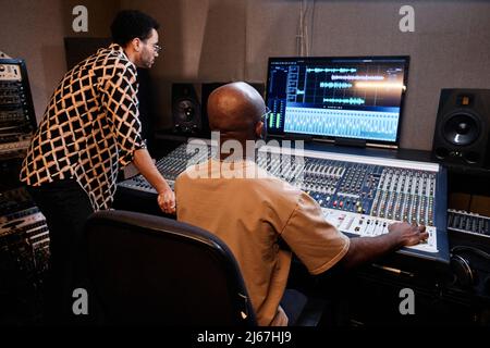 Reifer afroamerikanischer Produzent und junger Sänger, der im Aufnahmestudio mit einem Mischpult Musiktitel kreiert Stockfoto