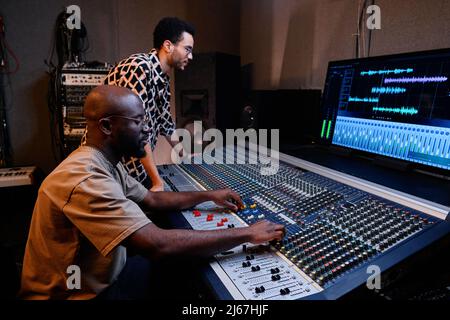 Professioneller reifer afroamerikanischer Produzent und junger Musiker, der im Aufnahmestudio Track kreiert Stockfoto