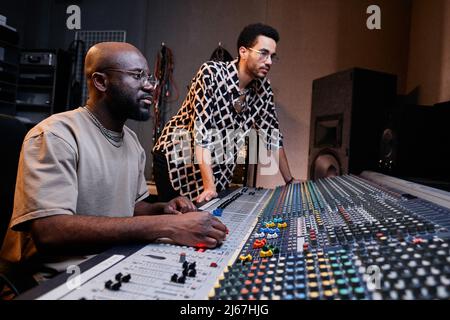 Moderner reifer Black Sound Engineer und stylischer junger Sänger, der im Aufnahmestudio an Musikstücken arbeitet Stockfoto