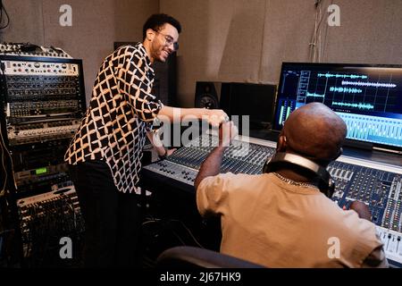 Erfolgreicher afroamerikanischer Musikproduzent und junger Sänger, der im Tonstudio die Arbeit an einem Song beendet und dabei Faustschlag macht Stockfoto