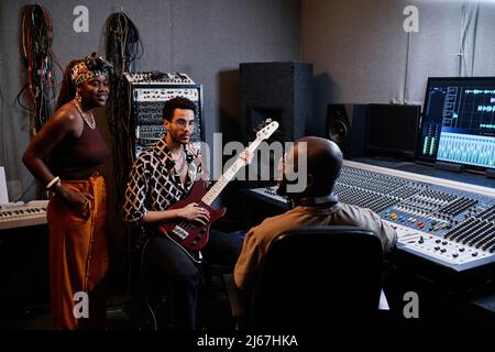 Reifer afroamerikanischer Musikproduzent und zwei junge Musiker diskutieren im Tonstudio etwas Stockfoto