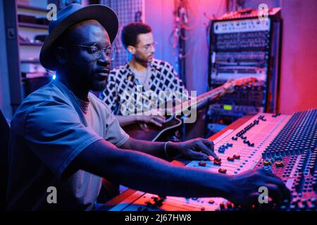 Selektive Fokussierung des stylischen, reifen afroamerikanischen Produzenten, der den Sound auf dem Mixer justiert, während ein junger Musiker Gitarre spielt Stockfoto