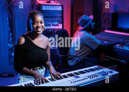 Fröhliche junge afroamerikanische Musikerin, die digitales Keyboard spielt, während die Produzentin am Mischpult im Aufnahmestudio arbeitet Stockfoto