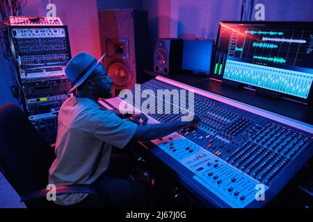 Stilvoller, reifer afroamerikanischer Mann mit Hut, der Soundtrack mit Mischpult im Aufnahmestudio in Neonlicht kreiert Stockfoto