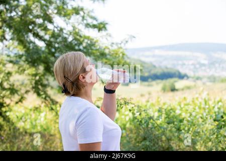 Profilfoto einer sportlich blonden Frau, die eine Digitaluhr an der Hand trägt und nach dem Joggen Wasser aus der Plastikflasche im Freien trinkt. Stockfoto