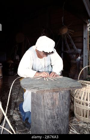 Williamsburg, VA. USA 9/1987. Korbweber. Master Korb Weber mit Holzstreifen in einem Korb von vielen Größen gewebt zu tragen oder zu speichern Gemüse, Maisähren, Obst, Eier, Nähgeräte, Feuerholz für die Küche, Feuerholz für den Herd und Waschlappen. Die Korbweberei ist 1000s Jahre alt. Stockfoto