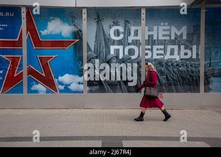 Moskau, Russland. 28.. April 2022 Eine Frau geht am Geschäft der russischen Armee mit einem festlichen Banner für die Feier des Siegestages im Schaufenster im Zentrum von Moskau, Russland, vorbei. Auf dem Banner steht: „Happy Victory Day!“ Stockfoto