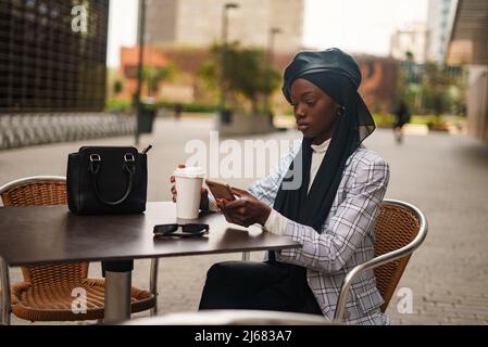 Ernsthafte schwarze Frau mit Smartphone am Tisch im Café im Freien Stockfoto