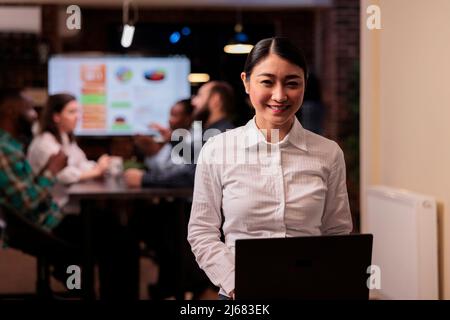 Porträt eines lächelnden asiatischen Geschäftsmitarbeiters, der in einem Startup-Büro einen Laptop hält und bei einem Treffen am späten Abend Überstunden arbeitet. Glückliche Frau im weißen Hemd posiert zuversichtlich mit Team macht Gruppenprojekt. Stockfoto