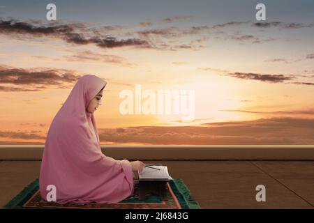 Asiatische muslimische Frau in einem Schleier sitzt und liest den Koran mit der dramatischen Szene Hintergrund Stockfoto