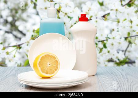 Plastikflaschen mit Spülmittel, Glas- und Fliesenreiniger, saubere Teller und eine Zitrone mit einem blühenden Kirschbaum auf dem Hintergrund. Waschen und c Stockfoto