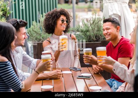 Eine multiethnische Gruppe von Freunden oder Studenten, die sich an einem Holztisch hinsetzen, während sie im Freien in einer Kneipe gemeinsam Bier toasten und trinken Stockfoto