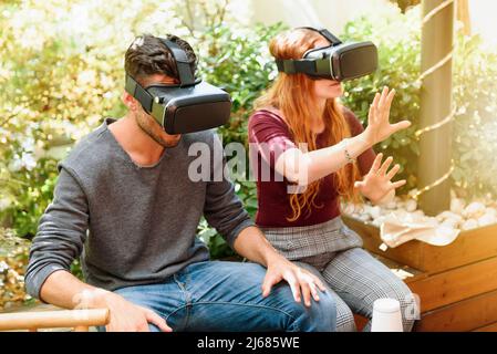 Fokussiertes Paar in legerer Kleidung, das auf einer Holzbank auf der Terrasse sitzt und an sonnigen Tagen VR-Visiere gegen grüne Büsche verwendet Stockfoto