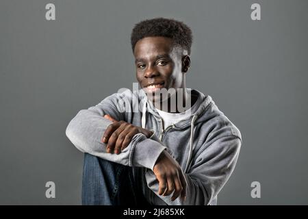 Positiver afroamerikanischer Mann, der lässige Kapuzenpullover und Jeans trägt und im Studio auf grauem Hintergrund sitzt und die Kamera anschaut Stockfoto