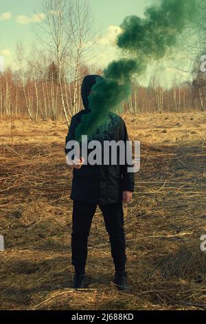 Ein Teenager in einer schwarzen Jacke und einer Maske hält eine grüne Rauchbombe Stockfoto