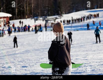 Umea, Norrland Schweden - 19. Februar 2022: Ein Mann hält sein Snowboard und steht im Hintergrund Schlange für einen Skilift Stockfoto