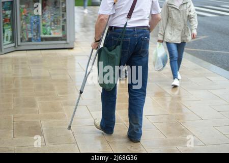 Ein Mann mit einer Krücke in der Stadt. Ein behinderter Mann geht die Straße hinunter. Der Typ mit dem verletzten Bein. Stockfoto