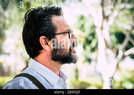 Seitenportrait des kaukasischen erwachsenen reifen Mannes mit Bart und Brillen Blick vor dem Park Natur geen defokussed Hintergrund. Ernst und Beruf Stockfoto