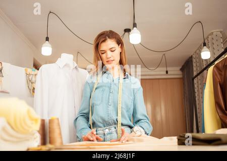 Porträt einer Näherin im Dachatelieratelieratelierin. Schönes Zimmer, Dummy mit weißem Hemd, Theke mit fertigen Kleidern. Kleines Unternehmen. Stoff Stockfoto