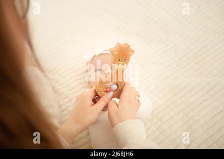 Kind liegt auf dem Bett, spielt mit Spielzeug, Mutter lehrt ihre Tochter, neugeborenes Baby im Schlafzimmer. Holzspielzeug für die Entwicklung. Stockfoto