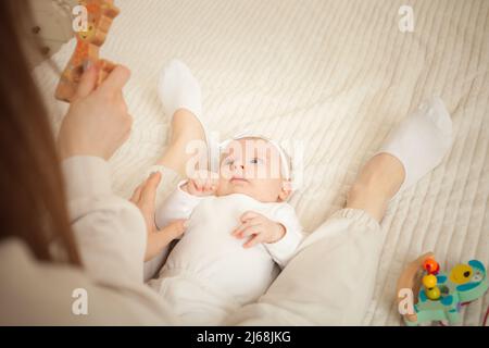 Kind liegt auf dem Bett, spielt mit Spielzeug, Mutter lehrt ihre Tochter, neugeborenes Baby im Schlafzimmer. Holzspielzeug für die Entwicklung. Spaß Stockfoto