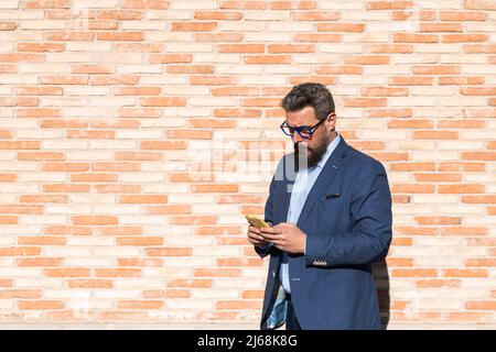 Geschäftsmann, der ein Smartphone benutzt, während er im Freien gegen eine Ziegelwand steht. Stockfoto