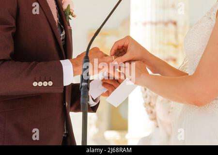 Die Hand der Braut legt den goldenen Ring an ihrem Hochzeitstag in einem Garten auf die Hand des Bräutigams. Stockfoto