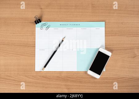 Leerer wöchentlicher Planner-Notice-Block auf einem Schreibtisch in einem Büro. Leerer Zeitplan und ein Bleistift. To-Do-Liste und Büroklammern auf dem Tisch. Smartphone liegt im Liegen. Stockfoto