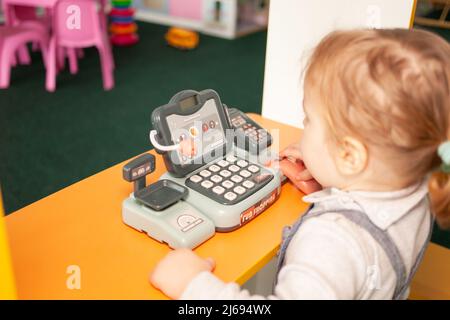 Ein kleines zweijähriges Mädchen spielt im Spielzimmer mit Spielzeug. Entwicklungszentrum, Kindergartentochter drückt die Tasten. Kasse für das Spielen im Laden Stockfoto