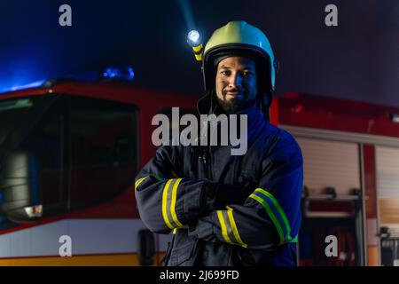 Porträt eines schmutzigen Feuerwehrmanns im Dienst mit einem Feuerwehrauto im Hintergrund in der Nacht, lächelnd. Stockfoto