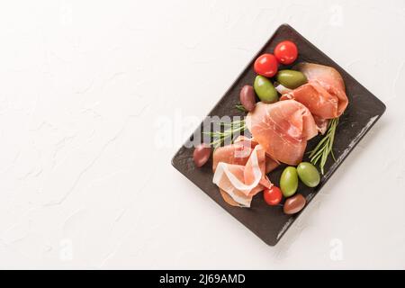 Draufsicht auf schwarzem Wurstbrett mit Proscuitto serviert mit Oliven auf weißem Beton-Tischhintergrund mit Kopierfläche. Schwarzer Teller mit Traditiona Stockfoto
