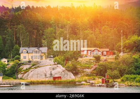 Schweden. Schöne Rote Und Gelbe Schwedische Holzhütten Häuser An Der Rocky Island Küste Im Sommerabend. See- oder Flusslandschaft. Sonnenuntergang Sommerhimmel Stockfoto