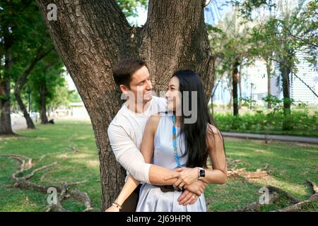 Nahaufnahme eines multiethnischen frisch verheirateten Paares, das sich neben einem Baum umarmt Stockfoto