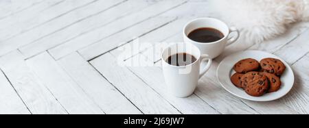 Frühstück am Morgen. Zwei weiße Tassen Kaffee und Schokoladenkekse stehen auf einem hellen Holzparkett und Kissen liegen in der Nähe. Gemütlichkeit, Tag Stockfoto
