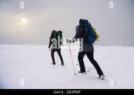 Die Jungs reden während der Winterexpedition. Sie tragen warme Jacken, Hüte und große Rucksäcke. Sie stehen im Schnee und halten Trekkingstöcke Stockfoto