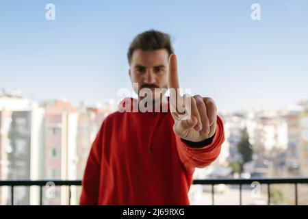 Junger ernsthafter Mann mit rotem Hoodie, der mit dem Finger nach oben zeigt und wütend ist, ohne Geste. Selektiver Fokus auf seine Hand. Stockfoto