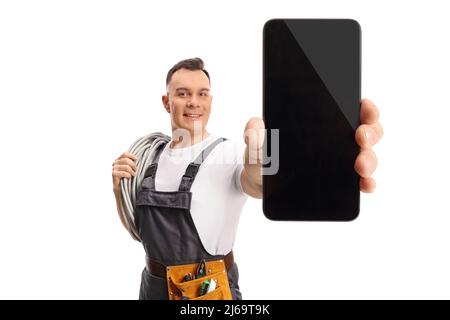Elektriker, der Kabel auf der Schulter trägt und ein isoliertes Smartphone auf weißem Hintergrund zeigt Stockfoto