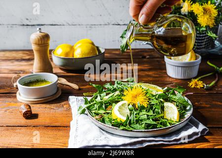 Dandelionensalat mit Olivenöl, Zitronensaft und Gewürzen auf braunem Holztisch. Menschliche Hand gießt Olivenöl zu Gericht. Stockfoto