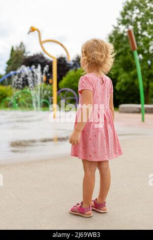 Kleines Mädchen, das im Sommer auf dem Spielplatz im Park spielt. Kleines Kind von hinten in der Nähe von Brunnen Stockfoto