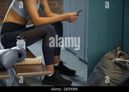 Unkenntliche Frau in Sportkleidung, die mit ihrem Smartphone auf der Bank im Umkleideraum im Fitnessstudio sitzt Stockfoto