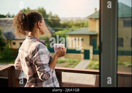 Rückansicht einer hübschen Frau in karierten Freizeitkleidung, die Kaffee trinkt und vom Balkon aus die benachbarten Wohngebäude und die ländliche Landschaft bewundert Stockfoto