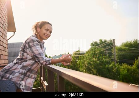 Schöne entzückende junge hispanische lockige haarige Frau in legerer Kleidung trinkt Kaffee und genießt den Blick auf die Landhäuser vom Balkon auf einem Stockfoto