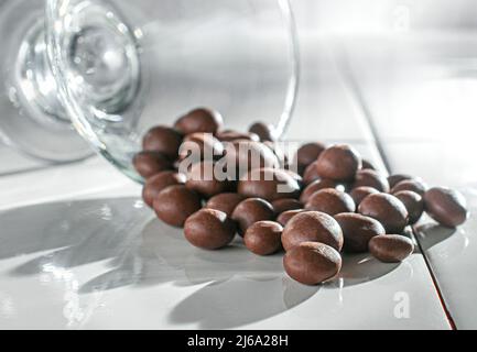 Mit Schokolade überzogene Rosinen, Haselnüsse, Kaffeebohnen und Mandeln, die aus einer Glastasse auf einem weißen Tisch mit Sonneneinstrahlung durch das Fenster gegossen werden. Stockfoto