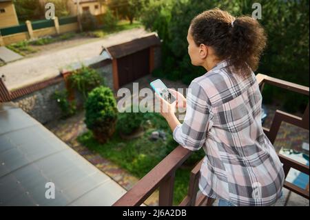 Blick von hinten auf eine junge dunkelhaarige Frau in legerer Kleidung, die auf dem Balkon eines privaten Kinderbetts mit dem Mobiltelefon unterwegs ist, im Internet surfen oder surfen kann Stockfoto