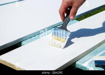 Männlicher Baumeister malt weiße Bretter für den Bau. Die männliche Hand hält die Bürste. Lackarbeiten, Hausrenovierung. Stockfoto
