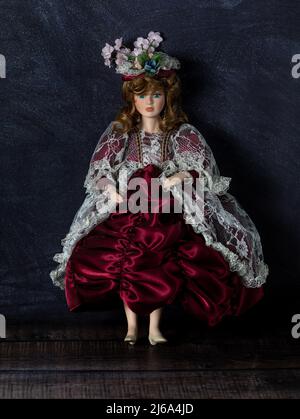 Erstaunlich realistisches Vintage-Spielzeug mit blauen Augen. Puppenkleid mit Blumen und Spitze. Selektiver Fokus. Kleid in leuchtendem Rot. Porzellan. Stockfoto