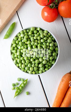 Frische, süße Erbsenschoten. Grüne Erbsen in weißer Schale mit frischen Schoten auf dem weißen Holztisch. Draufsicht. Stockfoto